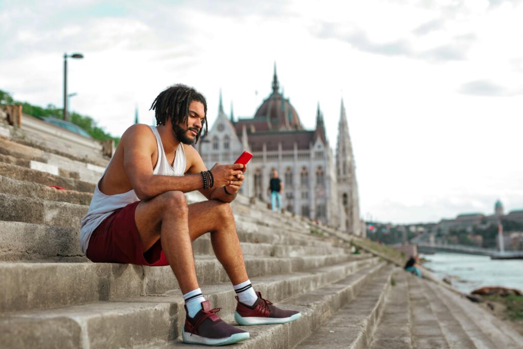 Young man sits casually on riverside steps in Budapest, engaged with his phone. Urban summer scene.